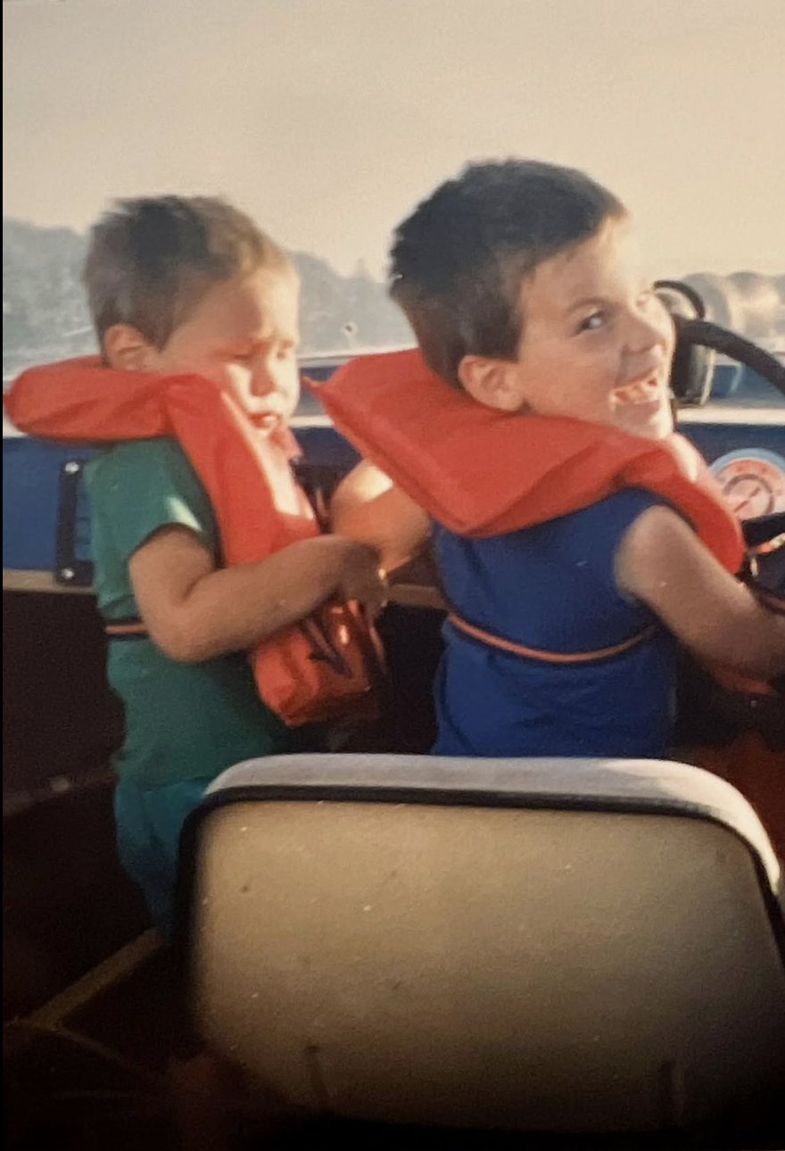 Matt and Mark Combe as young boys in orange life jackets aboard their grandfather's boat, Mark grinning at the helm