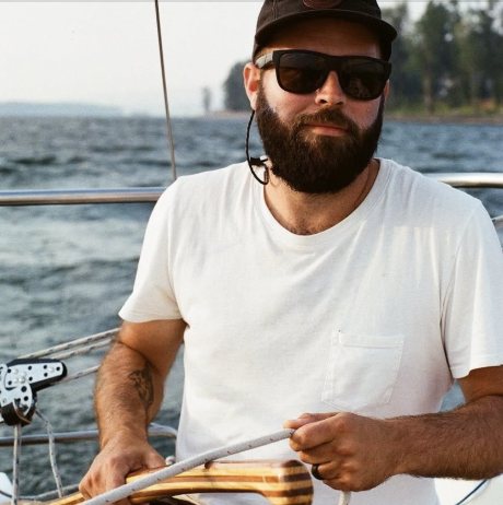 Captain Mark Combe at the helm under sail, in a white shirt, ball cap, and sunglasses