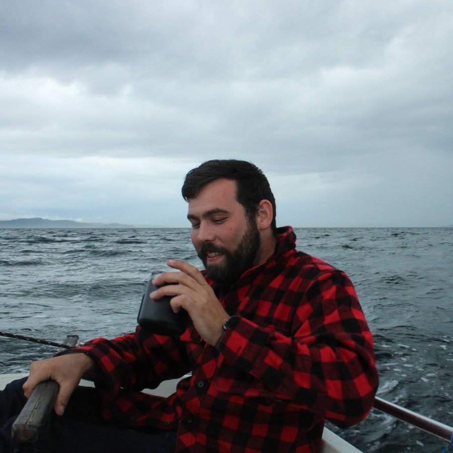 Matthew Combe in a red plaid flannel with his hand on the tiller, sailing across Puget Sound under a moody sky