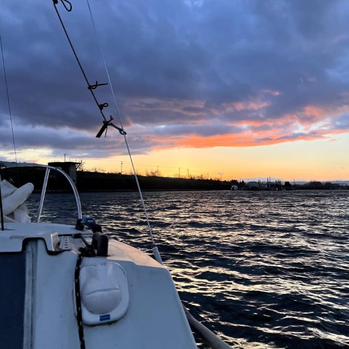 View from the foredeck of the sailboat at sunset, rigging in the foreground and an orange glow on the horizon over dark water